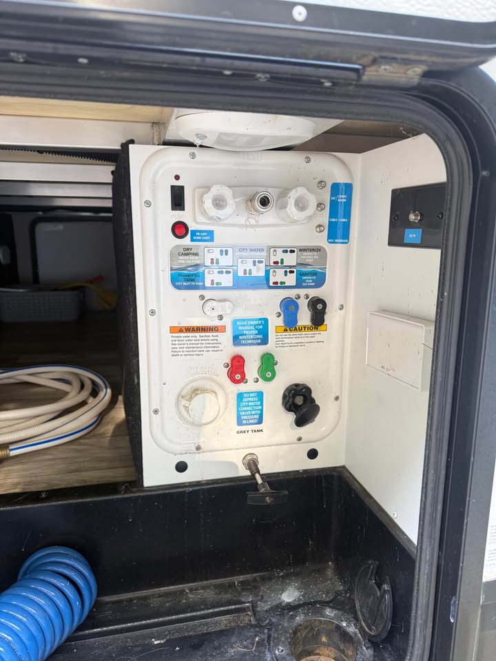 The 2021 Grad Design Reflection 297RSTS features a labeled RV water control panel with colored handles, warning labels, hose connections, and visible storage space inside an open compartment—perfect for organized setup and use.