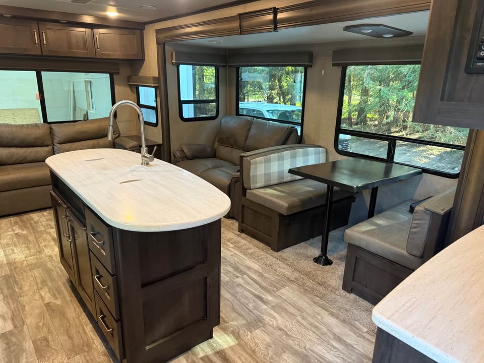 The 2021 Grad Design Reflection 297RSTS interior includes a kitchen island with sink, dark cabinets, sofa, cushioned dining booth with table, large windows showing trees and a car outside, and wood-look flooring.