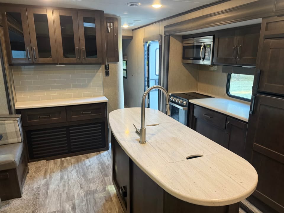 The 2021 Grad Design Reflection 297RSTS RV kitchen features dark wood cabinets, light countertops, a center island with sink, microwave, stove, refrigerator, tiled backsplash, laminate flooring, and abundant storage.
