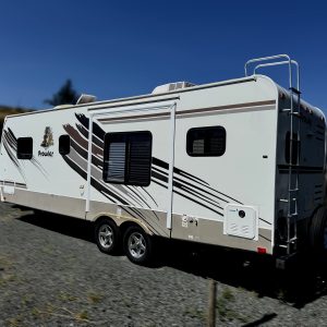 A 2007 - 26FT Prowler Luxury Lite travel trailer, white with black and brown stripes, is parked on gravel under a clear blue sky. Blurred trees and other RVs can be seen in the background.