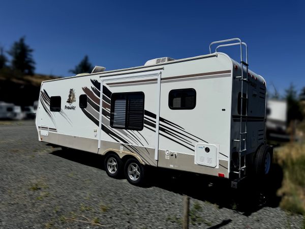 A 2007 - 26FT Prowler Luxury Lite travel trailer, white with black and brown stripes, is parked on gravel under a clear blue sky. Blurred trees and other RVs can be seen in the background.