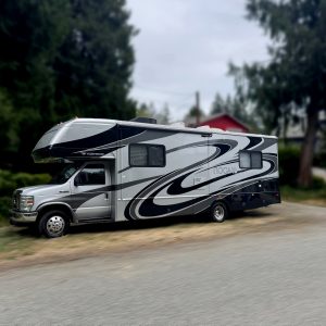 A silver and black 2007 Fleetwood Tioga 29V Class C motorhome is parked on the side of a rural road, with trees and a red house in the background.