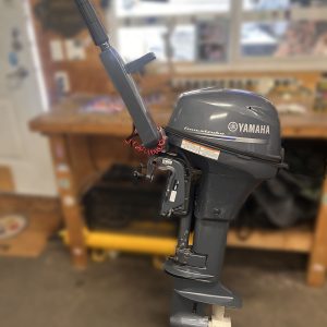 A 2014 Yamaha 9.9 HP Outboard High Thrust stands upright in a workshop, with a workbench and tools nearby and a red safety lanyard attached to its handle.