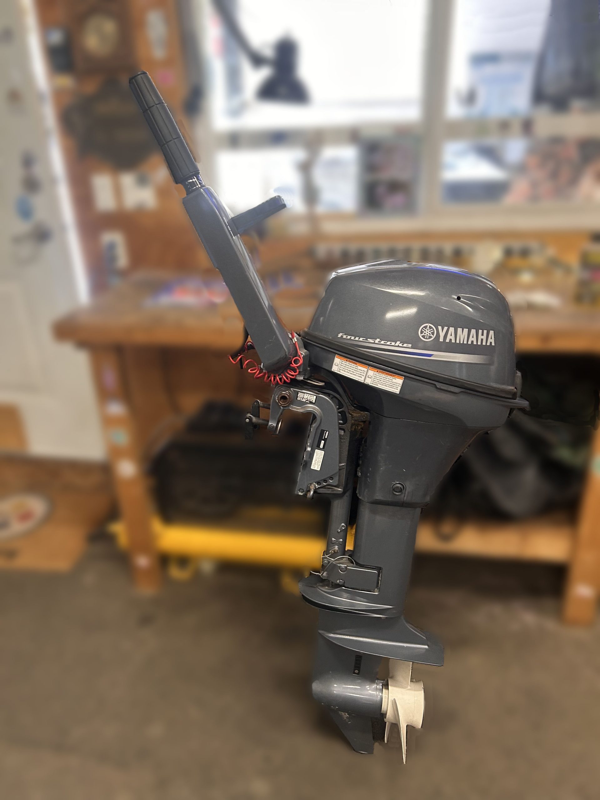 A 2014 Yamaha 9.9 HP Outboard High Thrust stands upright in a workshop, with a workbench and tools nearby and a red safety lanyard attached to its handle.