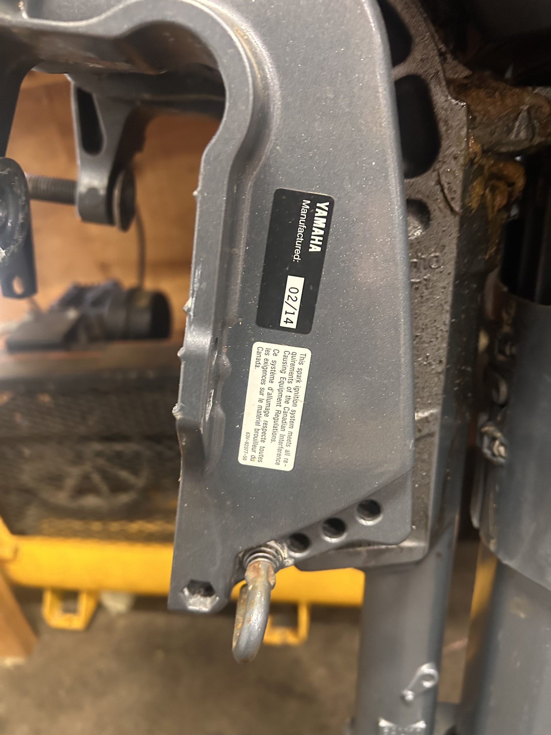 Close-up of a 2014 Yamaha 9.9 HP Outboard High Thrust mechanical part with a hook, showing Yamaha branding and labels against a blurred workshop background.