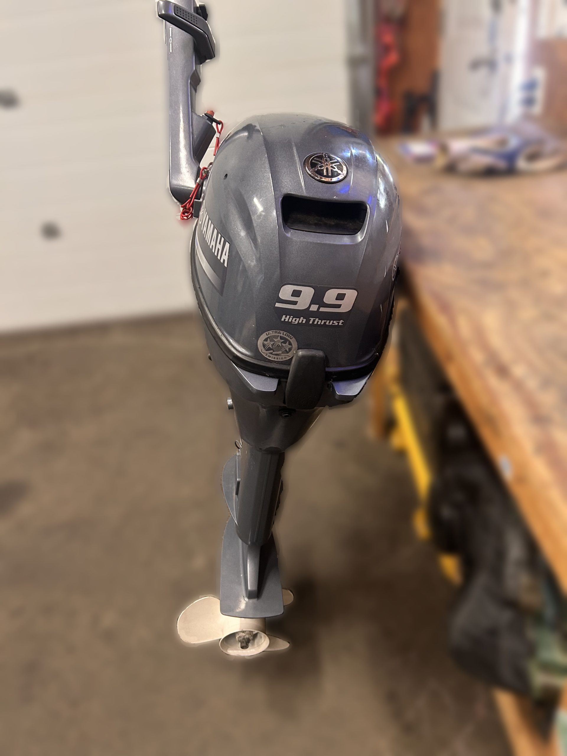 A 2014 Yamaha 9.9 HP Outboard High Thrust stands upright on a workshop floor beside a wooden workbench, with tools visible in the background.